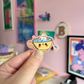 Bloemkolie Teatime Duck sticker: schattige witte eend in een lachende gele theekop met blauwe vijverthee en roze rand.