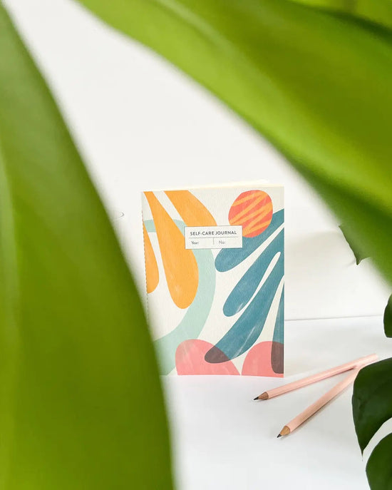 Fin Studio 'Nora' self-care notitieboek met abstracte blauwgroene en oranje vormen, groene bladeren en roze potloden.