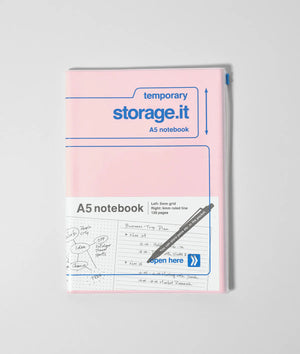 Roze A5 Storage.it notitieboek met blauwe tekst en een transparante hoes met ritssluiting tegen een minimalistische grijze achtergrond.