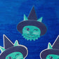 Knipperende groene katsticker met een zwarte heksenhoed en hoektanden op een glinsterende blauwe achtergrond.