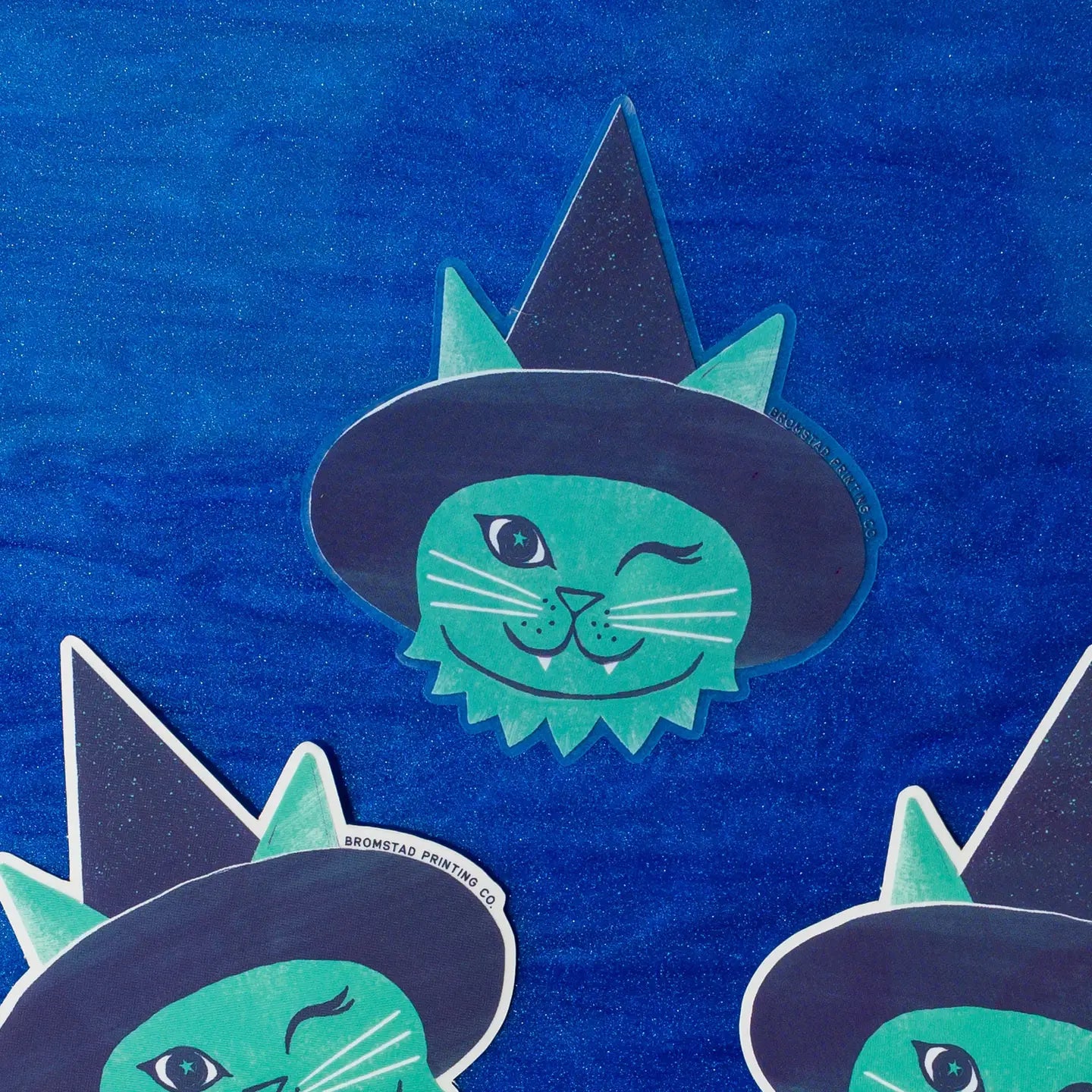 Knipperende groene katsticker met een zwarte heksenhoed en hoektanden op een glinsterende blauwe achtergrond.
