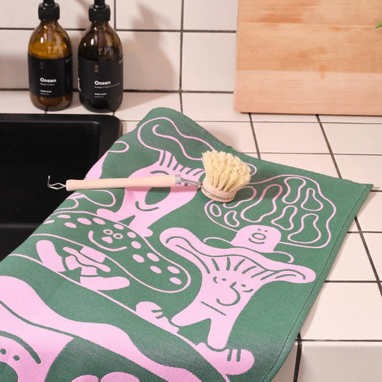 Groene en roze Wrap theedoek met grillige geïllustreerde paddenstoelkarakters op een betegeld aanrecht.