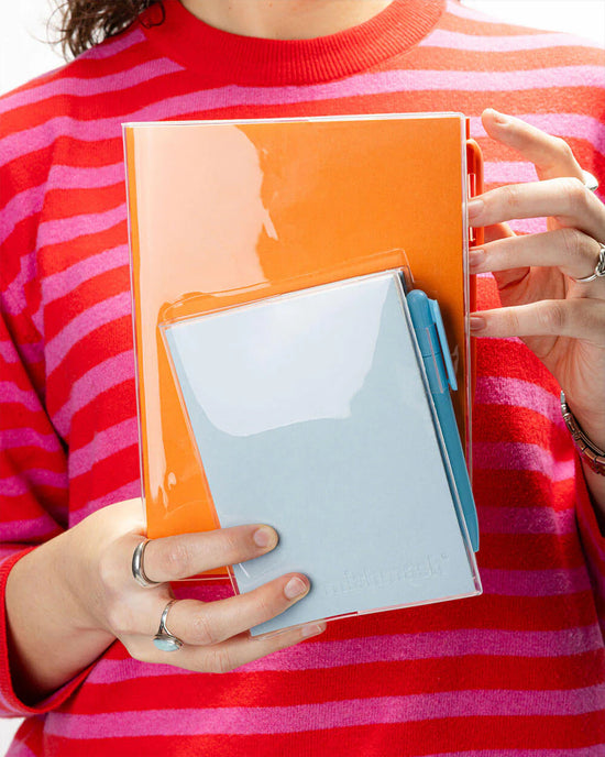 Transparante plastic A6 mishmash beschermhoes op een oranje notitieboek, met een vak voor een blauw notitieblok en pen.